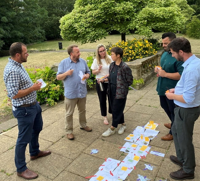 Group of men and women in a garden with paper and post-it notes having a discussion as part of Workshop 3.
